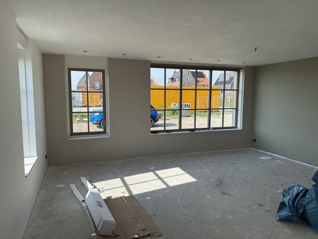Lege nieuwbouwwoonkamer met grijsgroene wanden en roedeverdeelde ramen, buiten een bouwkeet en huizen met pannendak